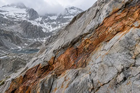 Broad orange-brown mineral seam running diagonally through fractured gray rock with a cloudy snowy basin beyond.
