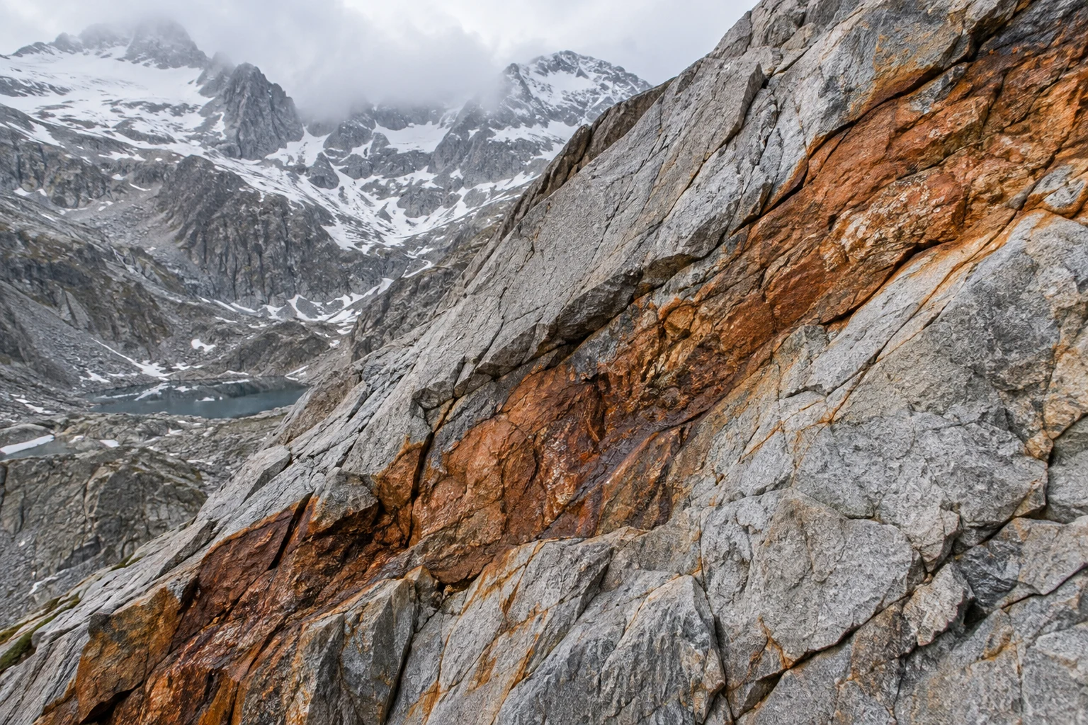 Broad orange-brown mineral seam running diagonally through fractured gray rock with a cloudy snowy basin beyond.