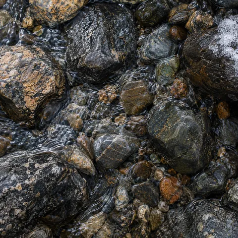 Overhead close view of varied brown and gray pebbles with thin flowing water, bright ripples and a small icy remnant at the upper right.