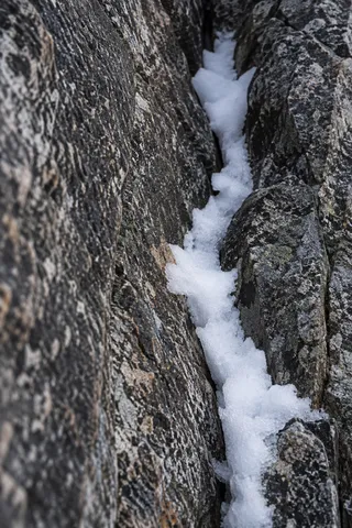 Close vertical crack between dark veined rocks filled with irregular granular white snow.