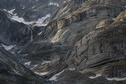Close gray mountain wall with curved rock layers, sheltered snow patches and thin descending meltwater streams.