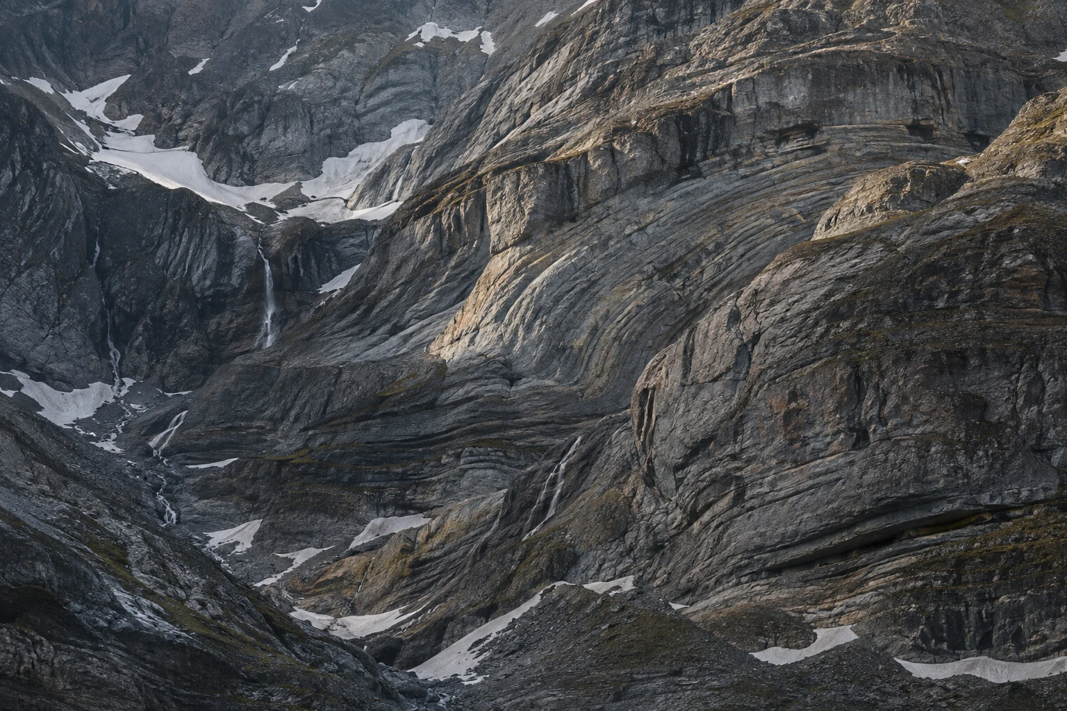 Close gray mountain wall with curved rock layers, sheltered snow patches and thin descending meltwater streams.