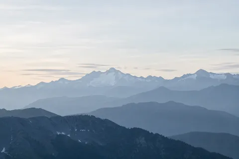 Successive blue mountain ridges fading into haze beneath a broad pale sky, with snow on the far range.