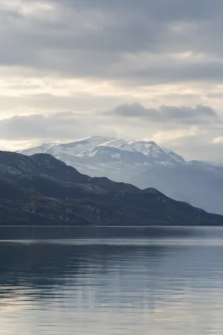 Pale snowy rounded mountain behind a dark autumn-speckled ridge, with a blue-gray lake and broad cloudy sky.