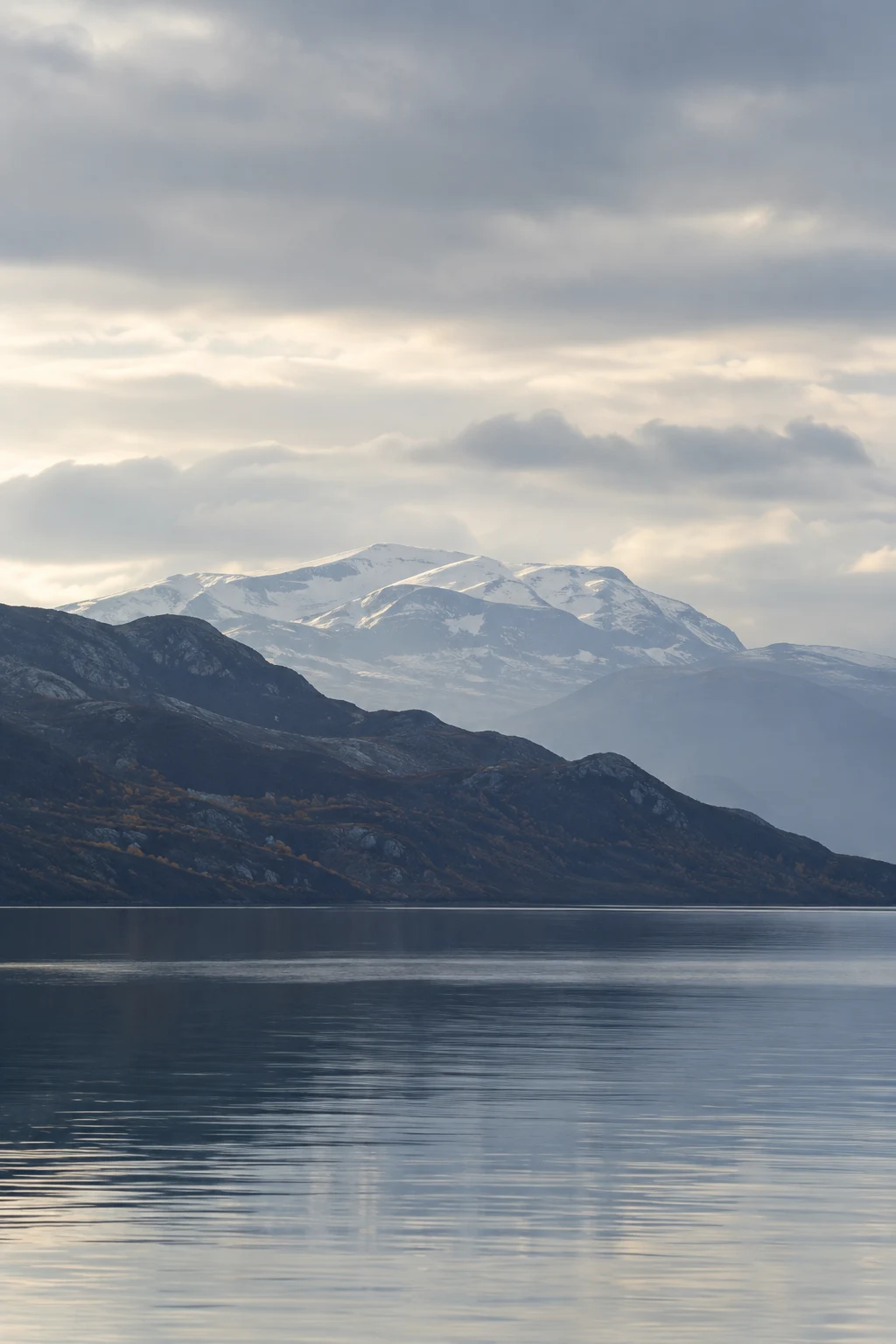 Pale snowy rounded mountain behind a dark autumn-speckled ridge, with a blue-gray lake and broad cloudy sky.