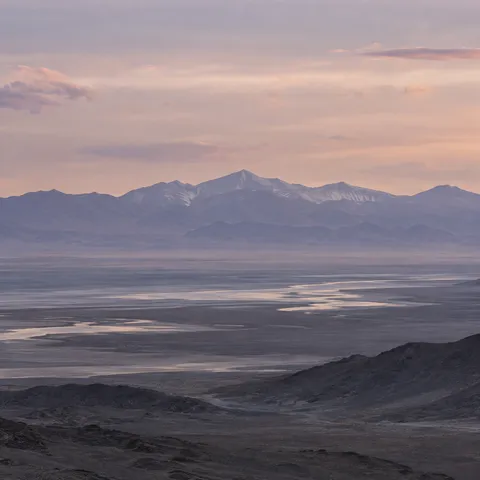 Distant violet-gray mountains above a hazy basin with pale winding water and a soft peach evening sky.