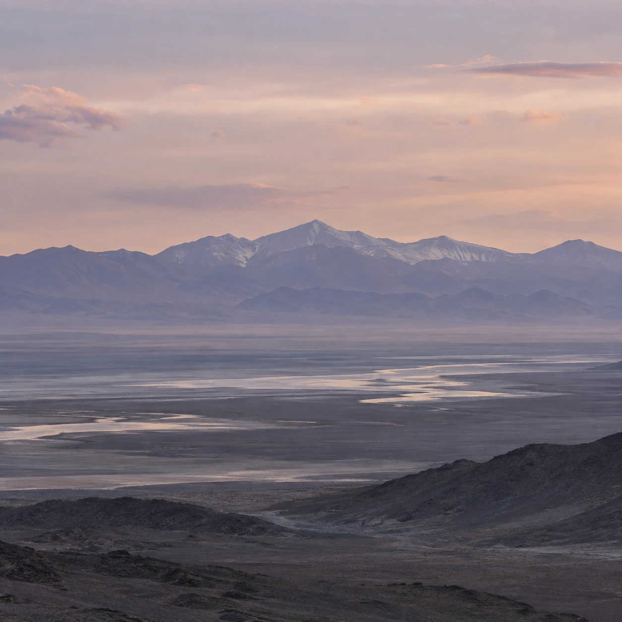 Distant violet-gray mountains above a hazy basin with pale winding water and a soft peach evening sky.