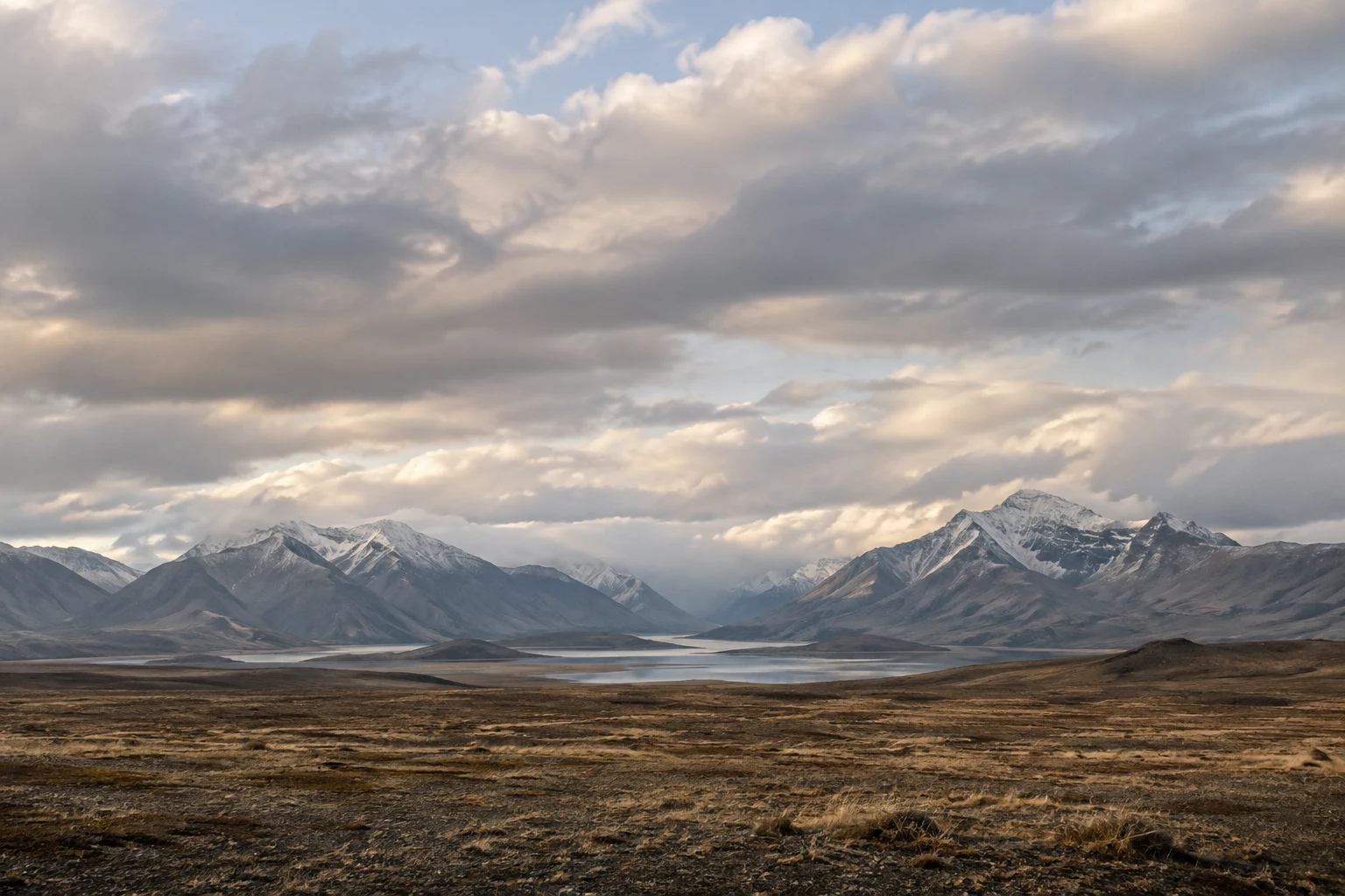 Broad low brown grassland before a thin lake and distant snow-touched mountains beneath a large cloudy sky.
