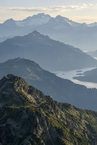 Portrait view of successive rocky and forested mountain spurs with a winding lake visible between them and a pale snowy range behind.