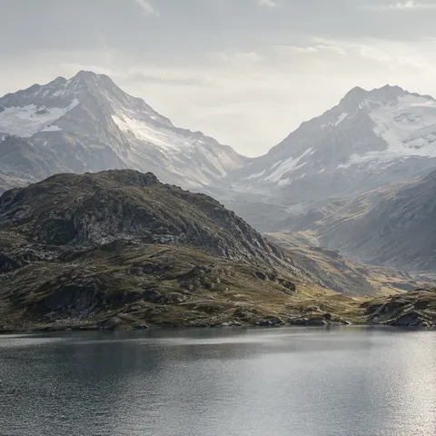 Two hazy snow-lined mountain slopes forming a saddle behind an olive rocky hill and finely rippled gray lake.
