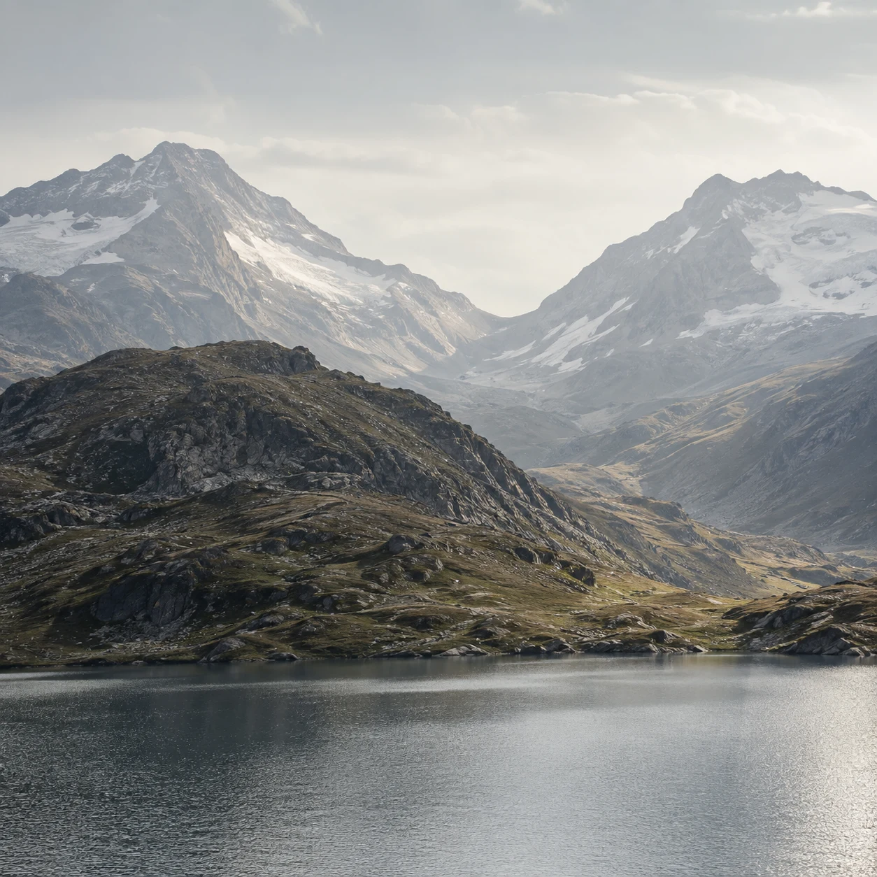 Two hazy snow-lined mountain slopes forming a saddle behind an olive rocky hill and finely rippled gray lake.