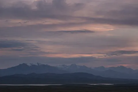 Low dark mountain range with scattered snow beneath layered mauve clouds and a thin strip of distant lake.