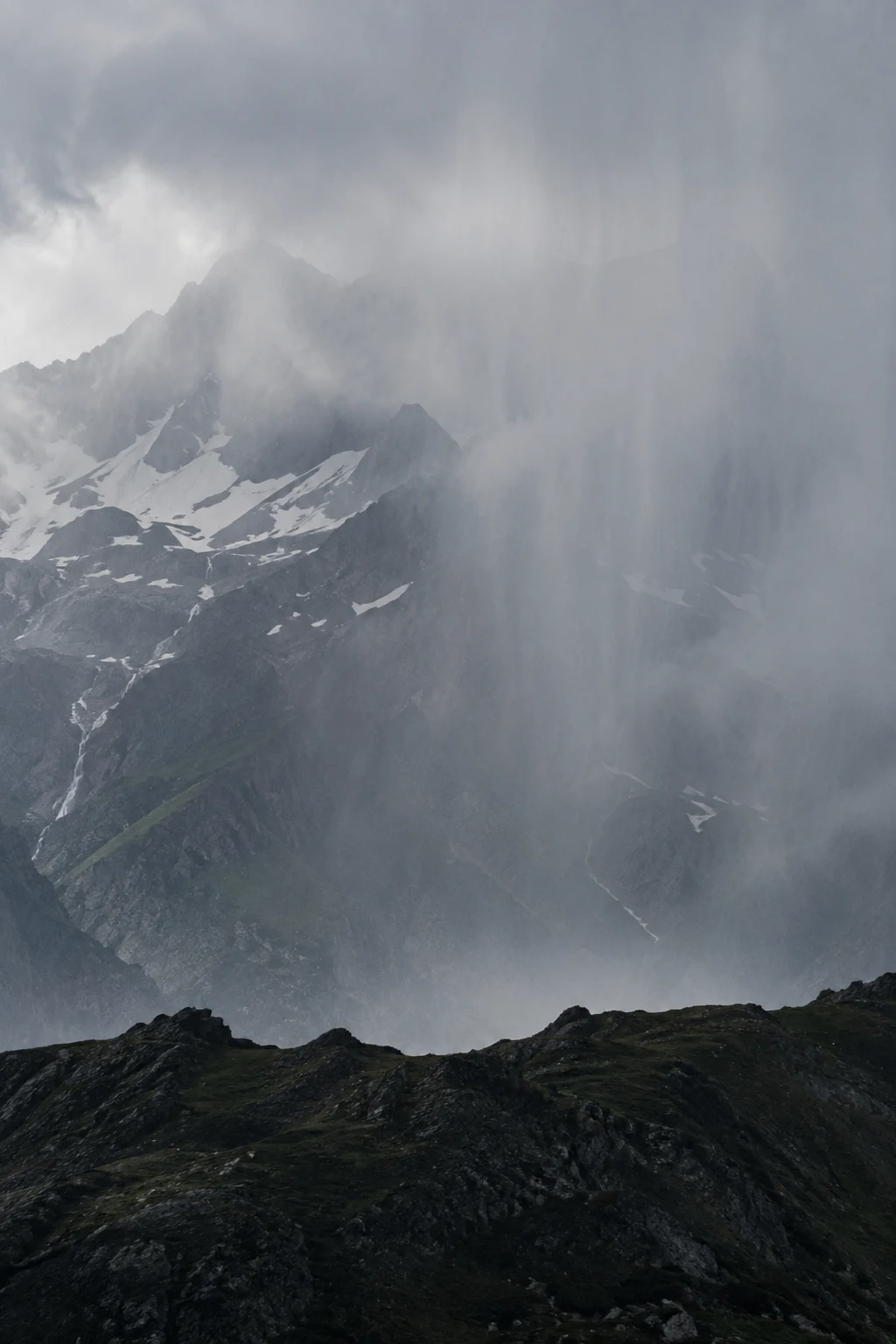 Portrait view of a gray snow-patched mountain partly obscured by descending rain and mist above a dark foreground ridge.
