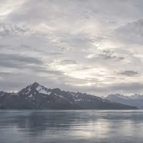 Low dark snow-marked mountain ridge at left across silver water, with a large softly glowing gray cloud sky.