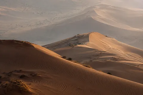 Layered ochre dunes with fine foreground ripples, sparse scrub and pale distant ridges fading into dusty air.