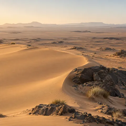 Curved warm sand dune wrapping around dark rocks and dry grass above a broad hazy desert plain.