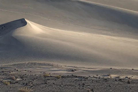 Long pale sand dune shoulder with a small left crest, fine ripples and a cool rocky foreground.