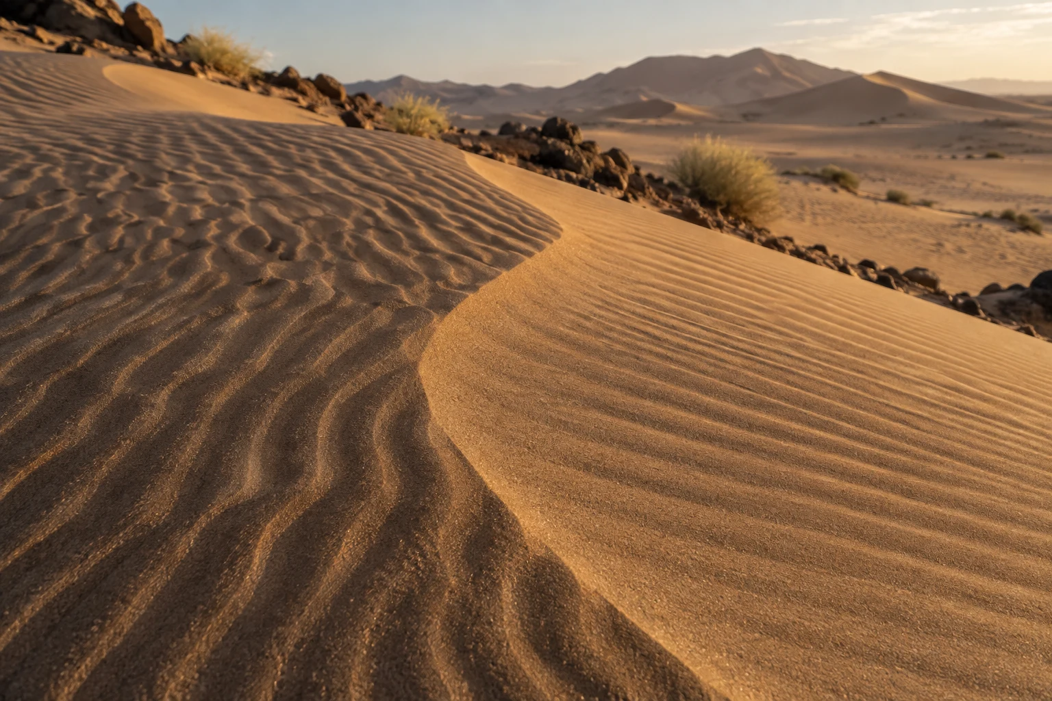 Two angled sand-ripple patterns meeting along a curved dune shoulder with a softly blurred desert background.
