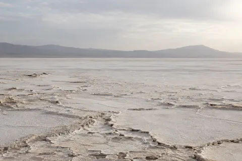 Wide pale salt basin with irregular crust plates in the foreground and a low hazy ridge beneath soft cloud.