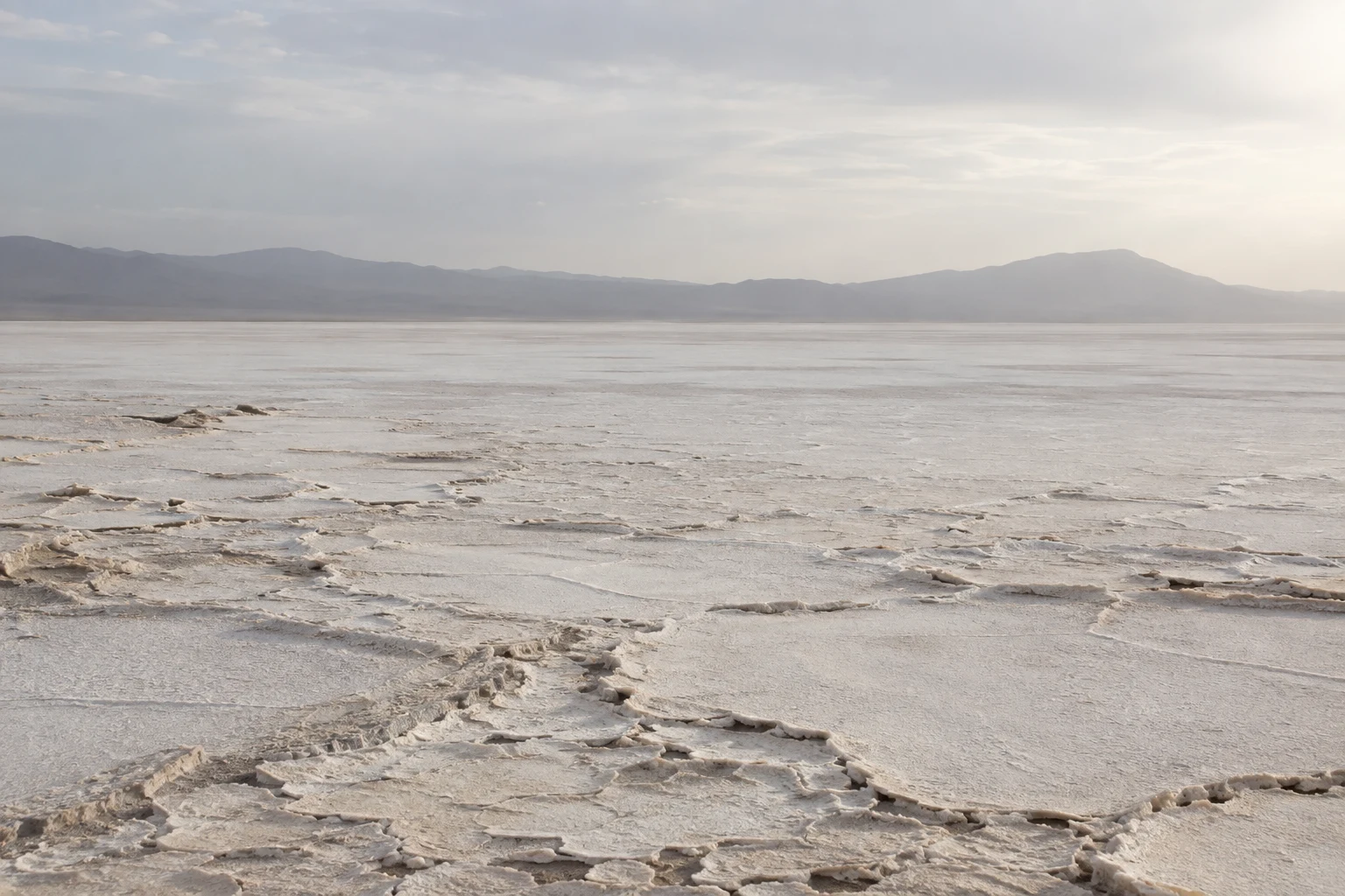 Wide pale salt basin with irregular crust plates in the foreground and a low hazy ridge beneath soft cloud.