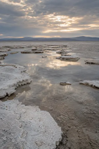Shallow reflective water between white salt-crust shelves beneath a softly lit cloudy sky and distant ridge.