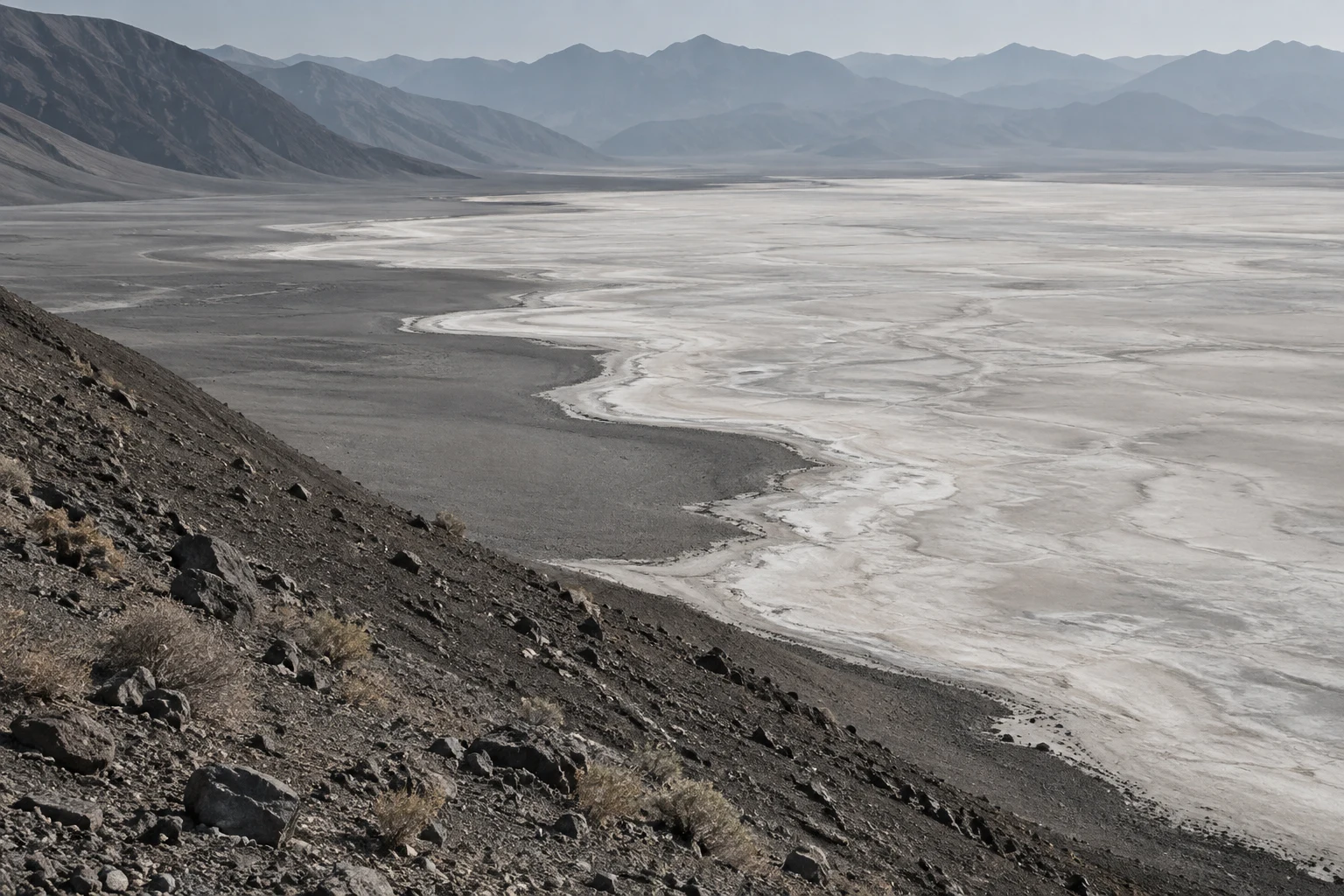 Pale salt-basin margin curving beside a dark gravel slope with stones and hazy ridges beyond.