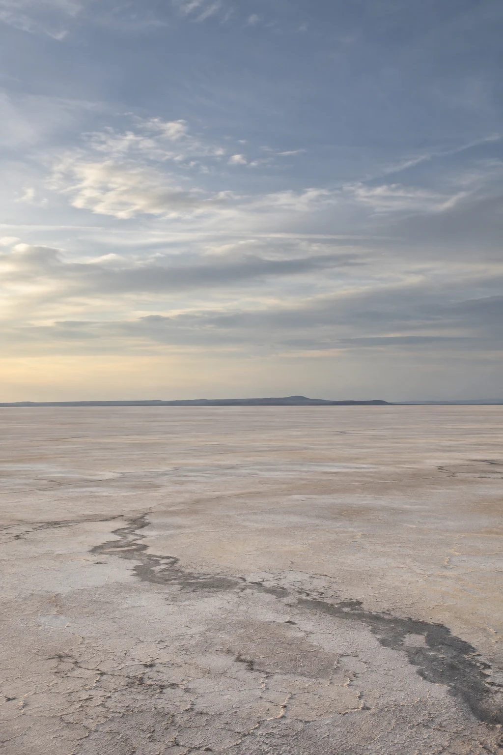 Pale flat salt basin with an irregular dark foreground seam beneath a broad blue-gray clouded sky.