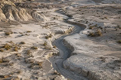 Narrow winding dry drainage channel cut into pale crusted earth with sparse scrub and gravel.