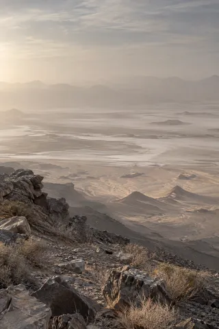 Pale desert basin veiled in dust beyond a rocky foreground ledge and dry scrub.