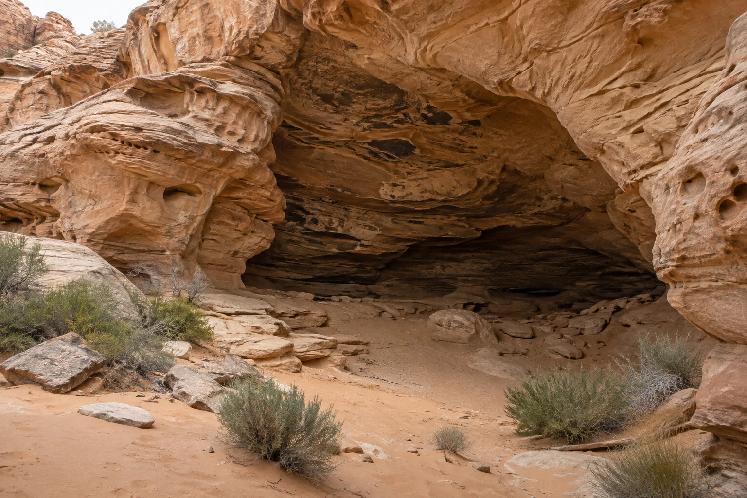 A shallow shaded sandstone alcove above sandy ground, loose rocks, and small desert shrubs.