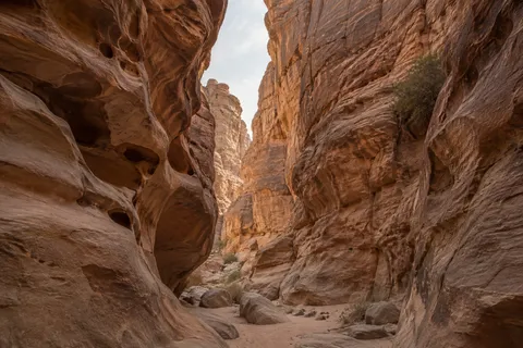 High sandstone walls, a narrow pale sky opening, and scattered boulders along a sandy canyon passage.