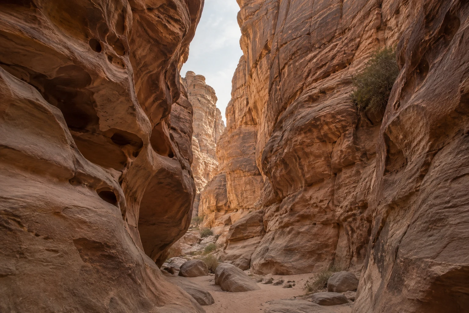 High sandstone walls, a narrow pale sky opening, and scattered boulders along a sandy canyon passage.