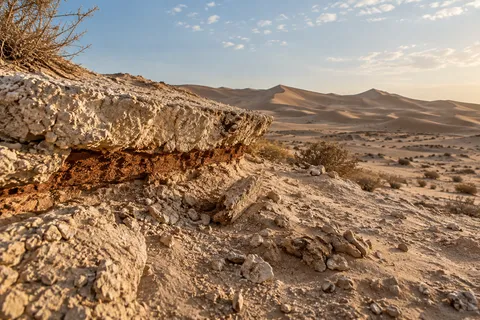 A rust-colored band beneath an ivory desert ledge with fallen fragments and distant sand dunes.