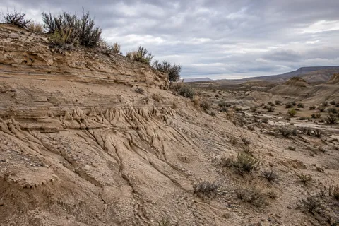 Erosion grooves beneath horizontal earth layers beside a dry shrub-covered basin under gray clouds.