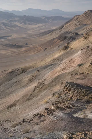 Muted gray, ochre, and dusty rose desert slopes descending toward a hazy basin and distant hills.