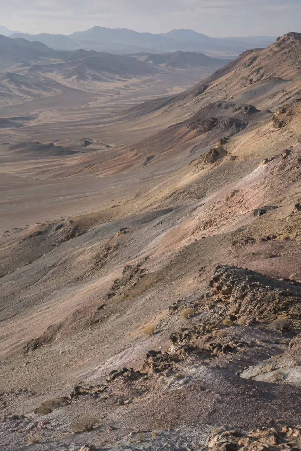 Muted gray, ochre, and dusty rose desert slopes descending toward a hazy basin and distant hills.