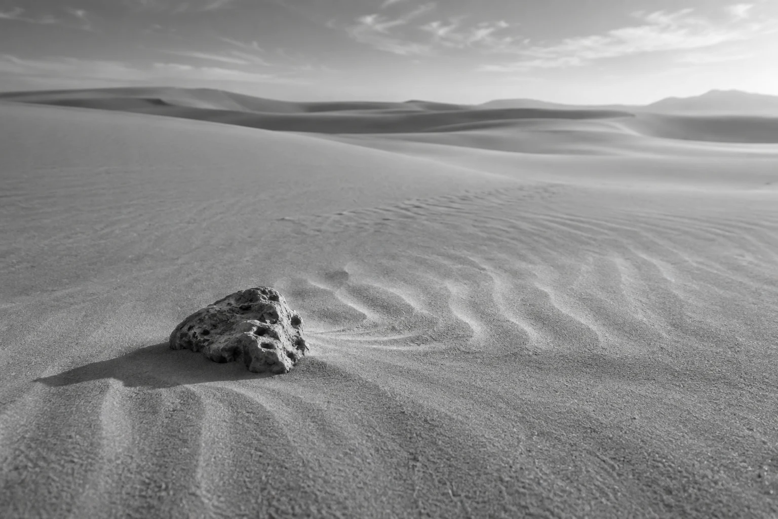 Black-and-white small weathered stone on pale rippled sand with soft distant dune contours.