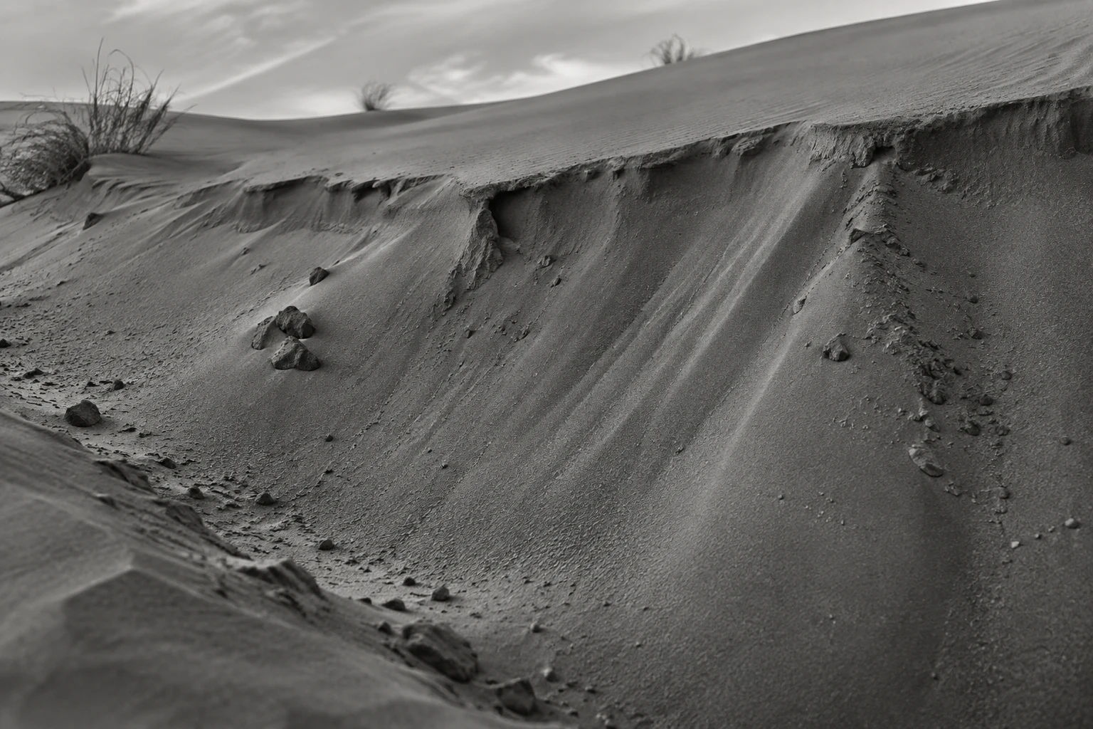 Black-and-white close view of a sandy slope below an uneven lip with small stones and sparse grass above.