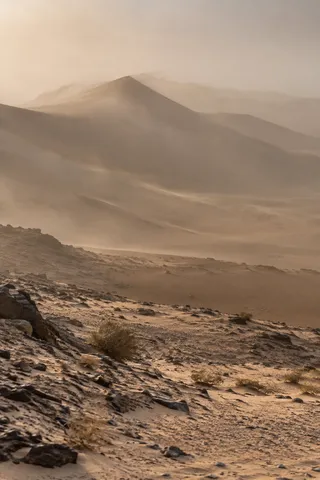 Distant dune crests softened by dusty haze above a rocky sandy foreground with sparse shrubs.