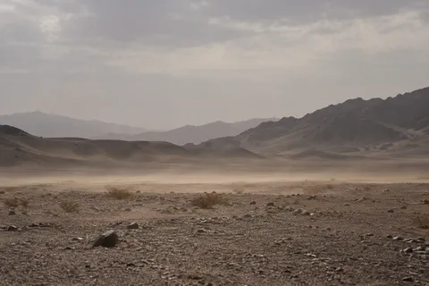 Low dust drifting across a gravel desert plain with dark stones, sparse shrubs, and layered hills.