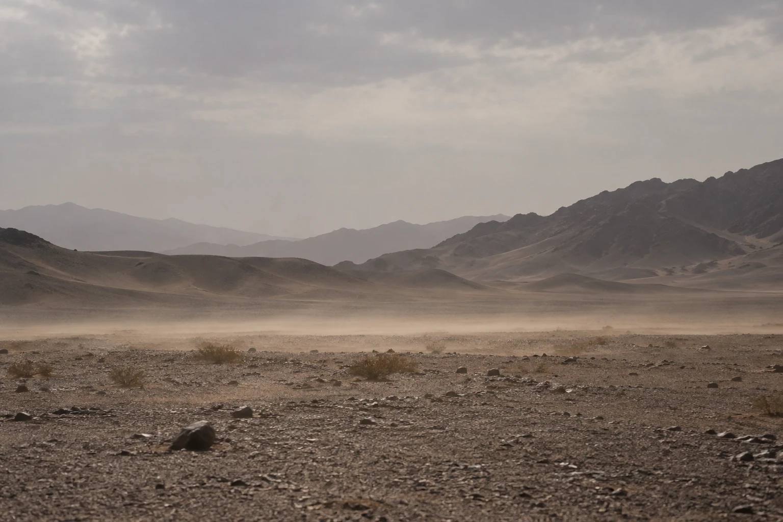 Low dust drifting across a gravel desert plain with dark stones, sparse shrubs, and layered hills.
