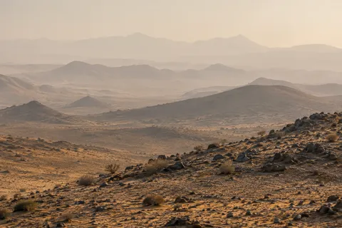 Overlapping low desert hills fading into warm dusty haze beyond a rocky shrub-covered foreground slope.