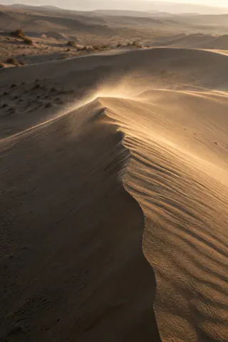 Fine sand blowing from an uneven dune crest in warm backlight above a dark slope.