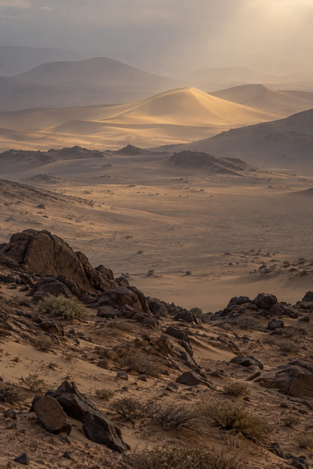 Distant dunes lit through haze beyond dark rocks and layered sandy terrain.