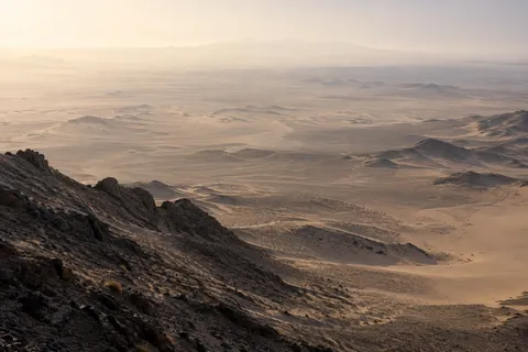 High view from a dark rocky ridge across a broad desert basin fading into pale warm haze.