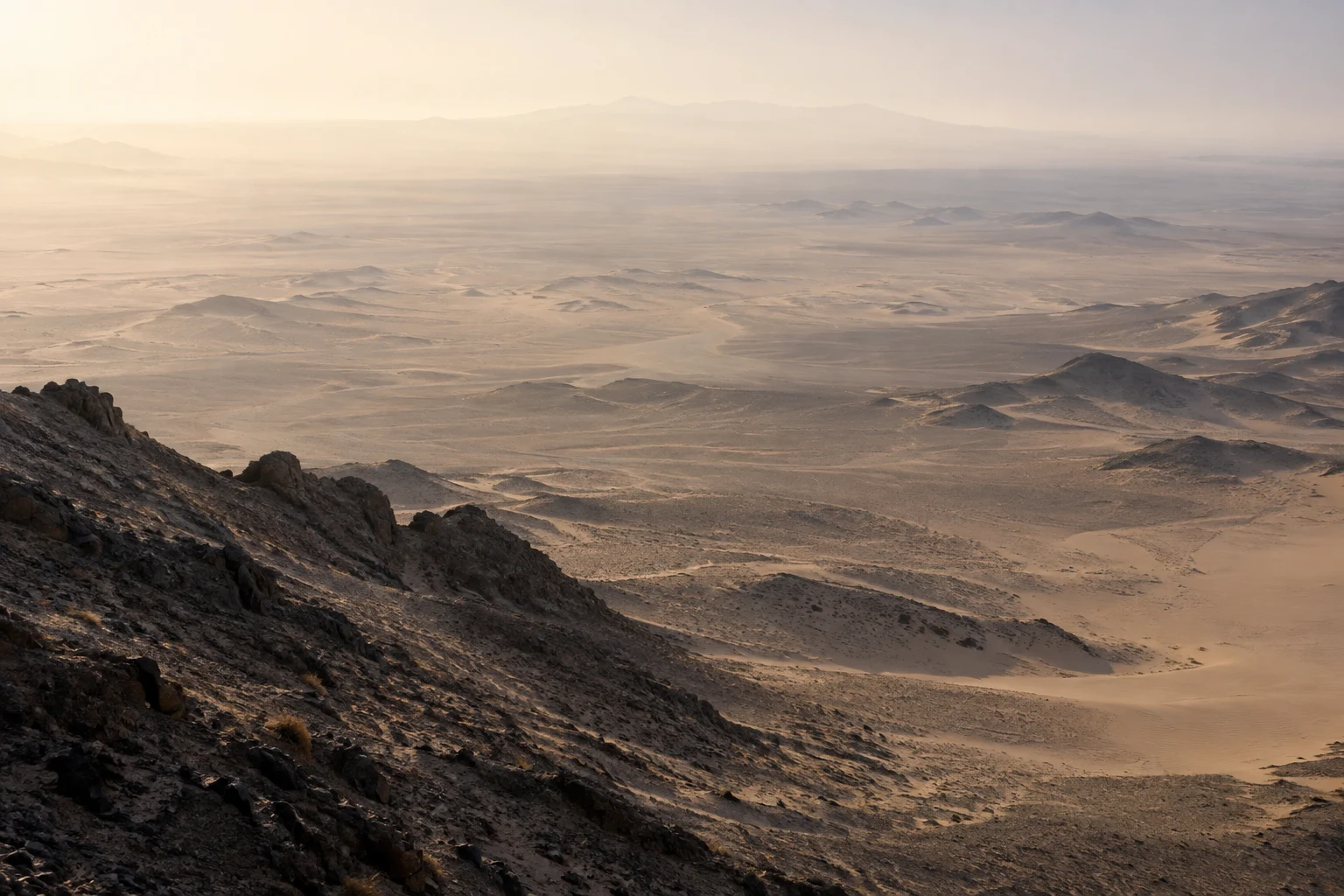 High view from a dark rocky ridge across a broad desert basin fading into pale warm haze.