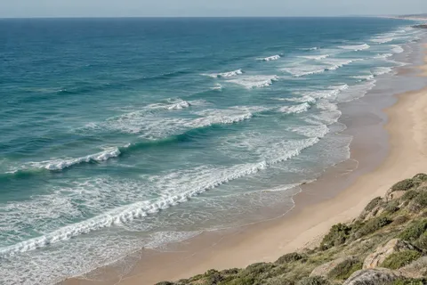 Long broken surf lines run beside an empty pale beach, viewed from a rocky coastal slope.