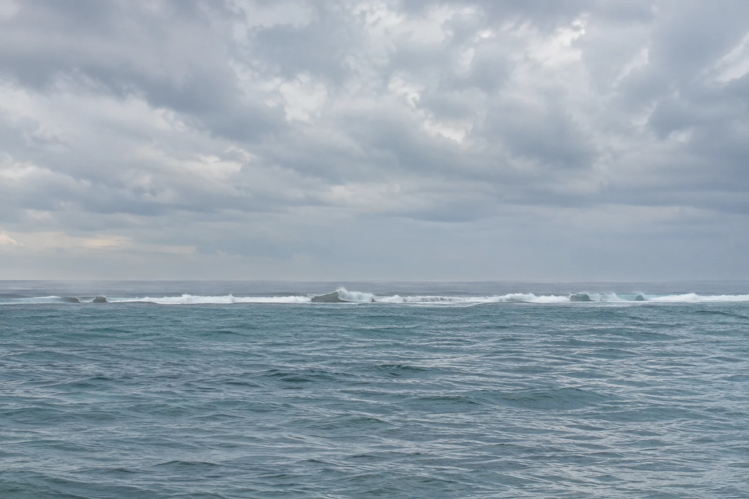 Low distant breakers cross a cool blue sea beneath a wide clouded sky.
