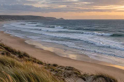Grasses on a sandy ridge overlook layered surf and a distant headland in evening light.