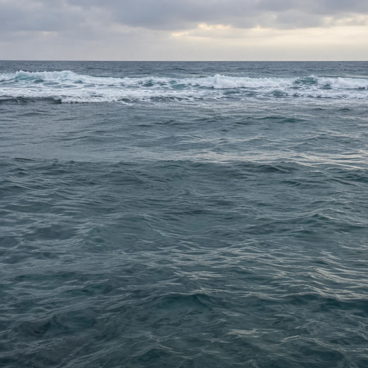 An uneven white breaker band crosses blue-gray water beneath a narrow clouded sky.